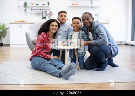 Ritratto completo di adulti multirazziali sorridenti e preschoolers coccolando intorno al tavolo da caffè in cucina open-space. Genitori amorosi e figli carini sentirsi felici mentre mettere blocchi in torre tumble. Foto Stock