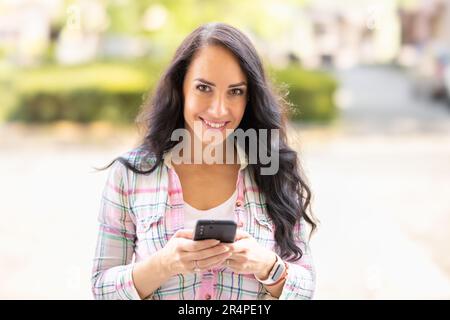 Una bruna sorridente sta scrivendo messaggi sul suo cellulare. Una giovane donna legge i messaggi ricevuti e scrive di nuovo su uno smartphone. Foto Stock