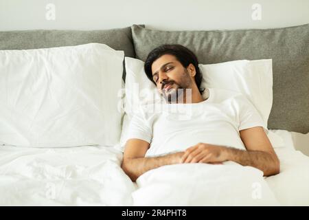 Un ragazzo medio orientale millenario che dorme, si trova su un letto bianco con spazio vuoto all'interno della camera da letto Foto Stock