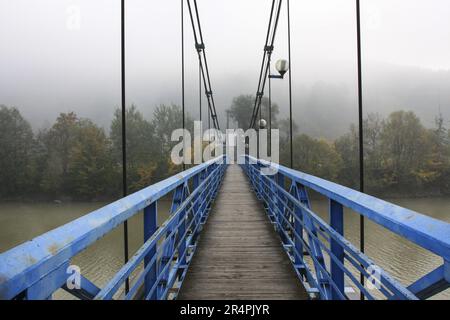 Ponte sospeso sul fiume sola, nebbia mattina d'autunno. Paesaggio mistico Foto Stock