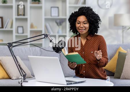 Una giovane giornalista afro-americana, blogger conduce un podcast, trasmesso a distanza da casa. Sedersi su un divano di fronte a un microfono e un computer portatile e leggere un libro. Foto Stock