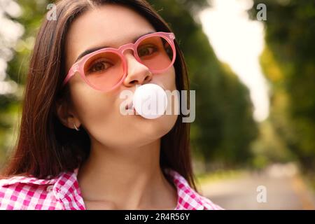 Bella giovane donna con occhiali da sole rosa che soffia gomma da masticare all'aperto Foto Stock