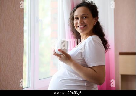 Donna incinta felice di etnia adulta in piedi alla finestra nella camera da letto di casa, sorride alla macchina fotografica, rilassarsi con una tazza di bevanda calda Foto Stock