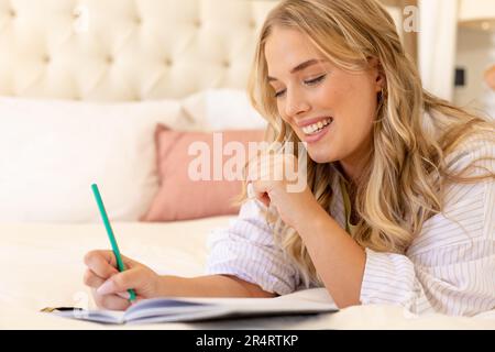 Giovane donna caucasica sorridente più grande con capelli biondi che scrivono nel libro mentre sdraiato sul letto Foto Stock