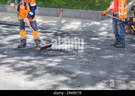 I lavoratori stanno riparando la strada, asfaltando la strada intorno ai tombini delle fognature e rimuovendo le macerie in eccesso con una spazzola e una pala. Foto Stock