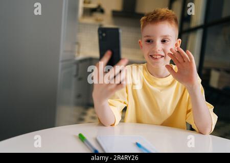 Ragazzo allegro che tiene lezioni di formazione a distanza tramite telefono cellulare, guarda una lezione online, videochiama in app e prende appunti Foto Stock