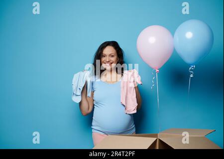 Bella donna in bruna incinta sorride, guardando la macchina fotografica, tiene neonati bodysuits su sfondo isolato blu Foto Stock
