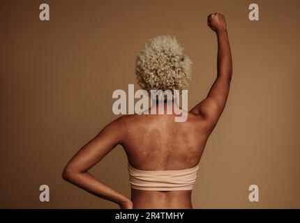 Donna con fiducia si alza in studio, la sua pelle marrone unica in bianco e nero con uno sfondo marrone. Gen Z femmina che alza il pugno per le donne empowerme Foto Stock