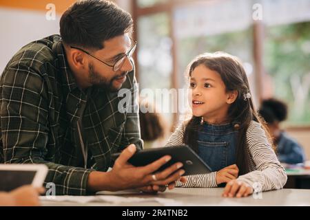 Insegnante di sesso maschile che parla con il suo studente mentre tiene un tablet digitale. Educatore della scuola elementare che imparte una lezione di alfabetizzazione digitale in una scuola elementare. Foto Stock