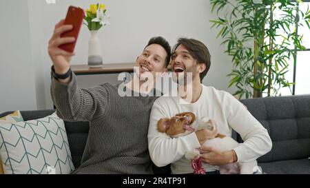 Due uomini si accoppiano facendo selfie da smartphone seduti sul divano con il cane a casa Foto Stock