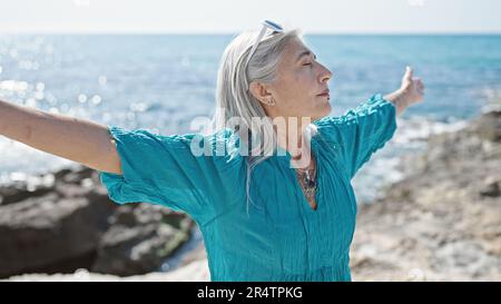 Donna di mezza età dai capelli grigi che respira con le braccia aperte al mare Foto Stock