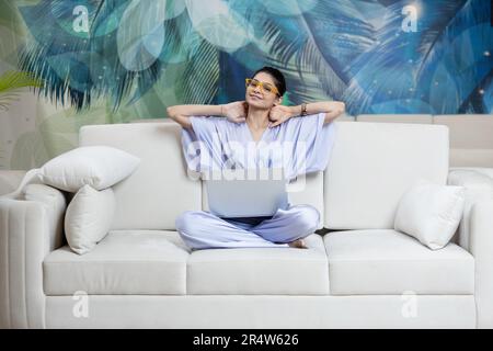 Immagine di una bella giovane donna rilassata che riposa mentre si siede con il computer portatile sul divano del soggiorno Foto Stock