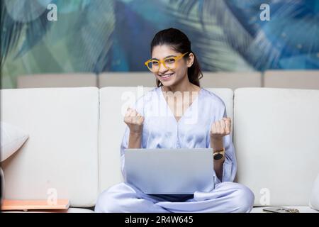 Giovane donna gioiosa seduta sul divano per celebrare il successo Foto Stock