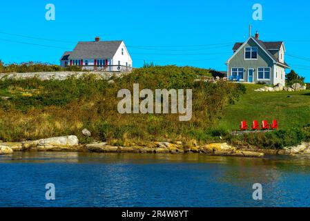 Due case colorate in legno con patio su una collina che si affaccia sull'oceano blu. Ci sono quattro sedie rosse Adirondack nel campo erboso vicino al mare. Foto Stock