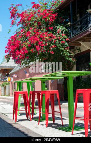 Tavoli e sedie in metallo color rosso vibrante e verde colorato, di dimensioni pub, si trovano su un marciapiede di fronte ad una caffetteria con alberi e fiori rossi fioriti Foto Stock