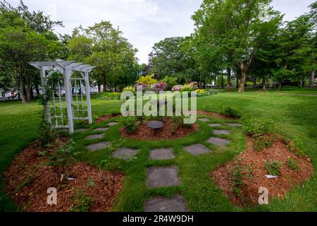 Un arco di legno bianco e pietre quadrate formano un cerchio in un giardino con alti e lussureggianti alberi di acero verde, cespugli di rose, piante, arbusti e erba verde. Foto Stock