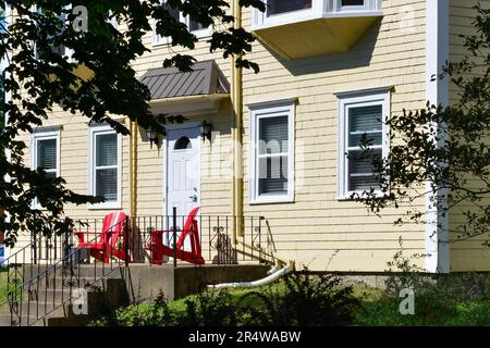 L'esterno di una casa in legno vintage di colore giallo clapboard stile country con sedie rosse Adirondack sul patio frontale. Foto Stock