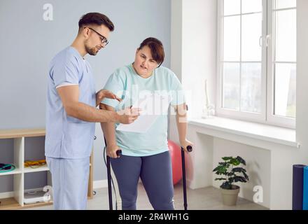 Una giovane infermiera che mostra una giovane paziente grassa con stampelle un referto che la aiuta a camminare. Foto Stock