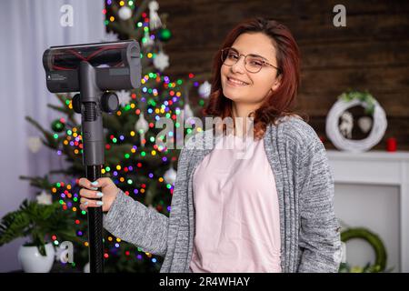 Una ragazza attraente tiene un aspirapolvere in mano contro lo sfondo di un albero di Natale. Foto Stock