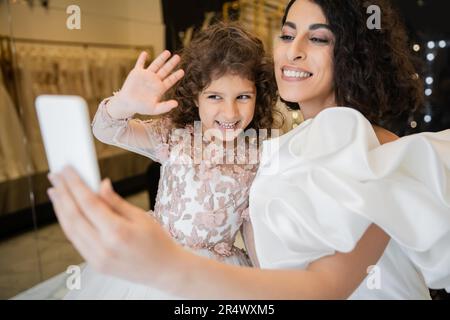 affascinante sposa mediorientale con capelli brunette in abito da sposa bianco con maniche in pelo e volant che tengono lo smartphone mentre felice figlia agita h Foto Stock