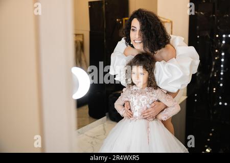 affascinante sposa mediorientale con capelli brunette in piedi in abito da sposa bianco con maniche e volant in pelo e guardando lo specchio mentre abbraccia la daug Foto Stock