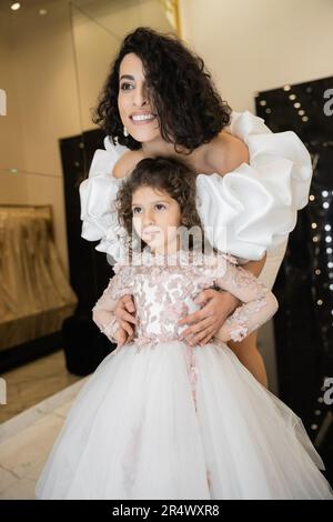 splendida sposa mediorientale con capelli brunette in piedi in bianco abito da sposa con maniche di puff e volant dietro la figlia carina e sorridendo insieme Foto Stock