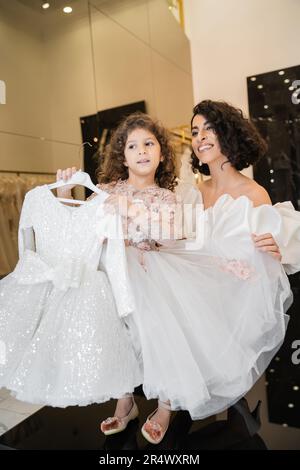 affascinante bride medio orientale con capelli brunette in piedi in abito da sposa bianco con maniche di puff e volant che tiene la gonna tulle della sua figlia carina Foto Stock