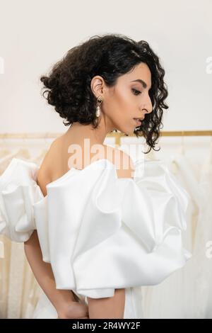brunette donna medio orientale con brunette e capelli ondulati posa in abito da sposa alla moda con maniche di puff e volant in boutique nuziale e look Foto Stock
