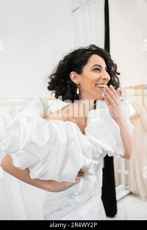 luminosa sposa mediorientale con bruna capelli ondulati ridere, coprendo la bocca e posando in abito da sposa alla moda con maniche di puff e volant in brid Foto Stock