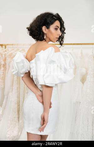 splendida sposa mediorientale con brunette e capelli ondulati in un elegante abito da sposa con maniche e volant in una boutique nuziale accanto a tul Foto Stock