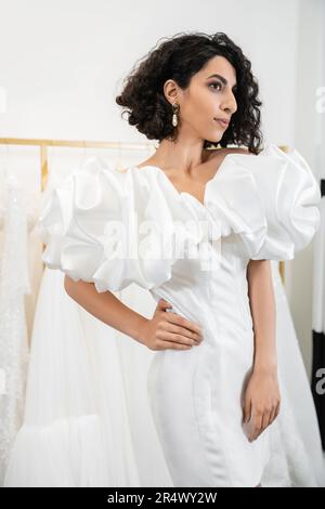 bruna sposa mediorientale con brunette e capelli ondulati che si posano a mano sul fianco in elegante abito da sposa con maniche in puff e volant in bou nuziale Foto Stock