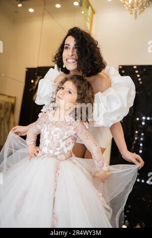 affascinante sposa mediorientale con capelli brunette in piedi in abito da sposa bianco con maniche e volant in pelo e guardare lontano mentre si tiene lo sci tulle Foto Stock