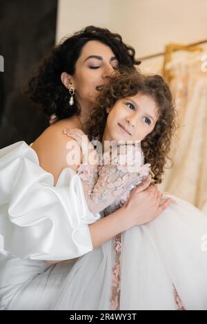 incantevole sposa mediorientale con capelli brunette e occhi chiusi in abito da sposa bianco con maniche in spugnetta e volant che abbraccia la figlia guardando lontano Foto Stock
