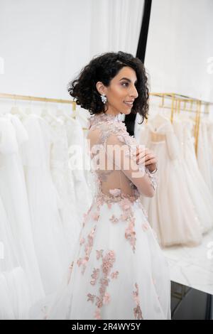 allegro medio orientale e donna brunette con capelli ondulati in piedi in uno splendido e floreale abito da sposa all'interno di un lussuoso salone nuziale intorno al bianco Foto Stock