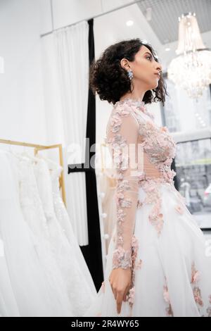 sposa-a-essere, splendida metà orientale e donna brunette con capelli ondulati in piedi in splendido e floreale abito da sposa all'interno di un lussuoso salone nuziale Foto Stock