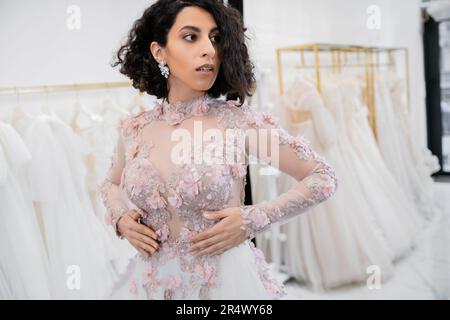 splendida sposa centro-orientale e brunette con capelli ondulati in piedi in uno splendido e floreale abbigliamento all'interno del lussuoso salone nuziale intorno tulle bianco f Foto Stock
