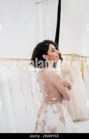 splendida donna centro-orientale e brunette con capelli ondulati in piedi in uno splendido e floreale abito da sposa all'interno di un lussuoso salone nuziale intorno al bianco Foto Stock