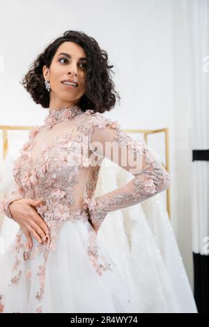 allegro medio orientale e donna brunette con capelli ondulati in piedi in elegante e floreale abito da sposa e guardando lontano all'interno del salone di lusso arou Foto Stock