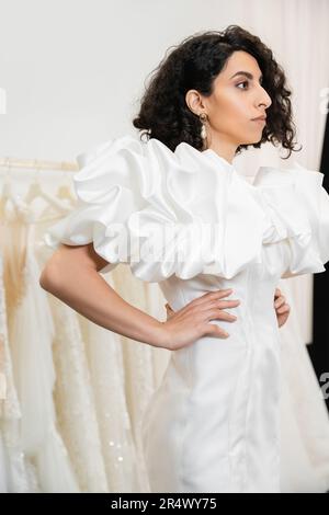 bruna sposa mediorientale con brunette e capelli ondulati che si posano con le mani sui fianchi in abito da sposa alla moda con maniche in puff e volant in bridal sa Foto Stock
