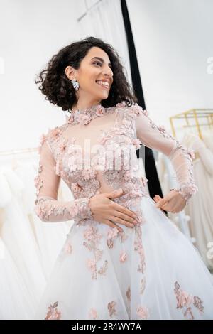 incantevole donna mediorientale e brunetta con capelli ondulati in piedi in stordimento e floreale abito da sposa e guardando lontano all'interno del salone di lusso a. Foto Stock
