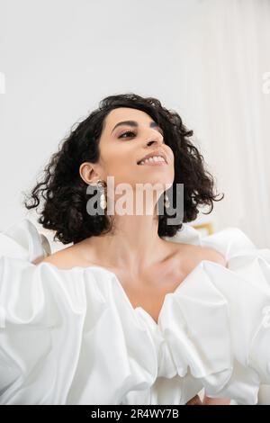ritratto di felice sposa mediorientale con brunette e capelli ondulati in posa in abito da sposa alla moda con maniche e volant in volant s Foto Stock
