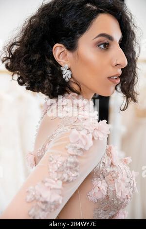 ritratto di deliziosa donna mediorientale con capelli ondulati in piedi in splendido e floreale abito da sposa e guardando lontano all'interno di un lussuoso salone ar Foto Stock