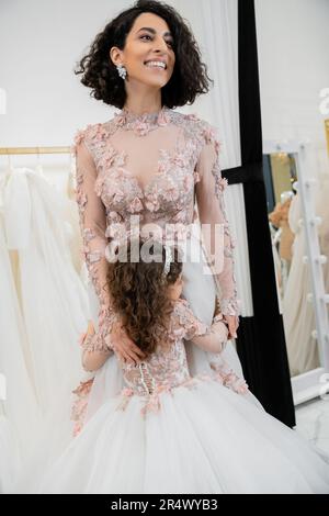 ragazza carina in abito floreale che abbraccia la madre allegra con capelli brunette in piedi in abito da sposa vicino sfocato abito bianco all'interno di luxurious sale nuziale Foto Stock