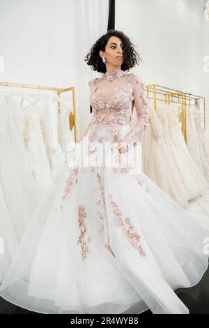 deliziosa e mediorientale donna con capelli ondulati cercando su elegante e floreale abito da sposa all'interno di luxurious salone nuziale, shopping, sposa-per-essere, Foto Stock