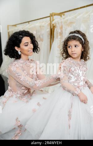 affascinante sposa medio orientale con capelli ondulati brunette in abito da sposa regolazione carino abito floreale di figlia in salone nuziale, shopping, speciale mome Foto Stock