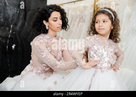 seducente sposa medio orientale con capelli ondulati brunette in abito da sposa regolazione carino abito floreale di figlia in salone nuziale, shopping, speciale mome Foto Stock