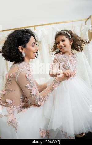 felice bride medio orientale con capelli ondulati brunette in abito da sposa floreale tenendo le mani con figlia sorridente in abito carino con gonna tulle in brid Foto Stock