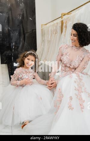 brunette medio orientale donna con capelli ondulati guardando ragazza allegra e sorridente vicino bianco abiti da sposa in salone nuziale, floreale, madre e daugh Foto Stock