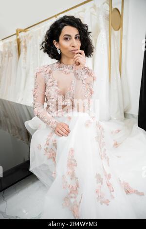 donna centro orientale brunette con capelli ondulati seduta in splendido e floreale abito da sposa vicino sfocato e abito bianco all'interno del lussuoso salone nuziale Foto Stock