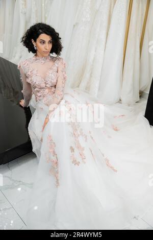 brunette medio orientale donna con capelli ondulati seduta in splendido e floreale abito da sposa con treno vicino sfocato e abiti bianchi all'interno di luxiou Foto Stock
